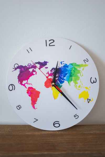 A large clock is resting on a wooden surface. The face of the clock has large numbers. The background is white and then the countries of the world are laid out in various colors.